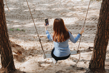 charming girl taking photo with smartphone while sitting on wooden swing in the forest near the wild sandy river beach