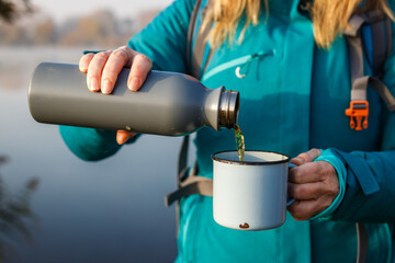 Woman is pouring hot drink from thermos into travel mug. Refreshment during hiking at lake in cold...