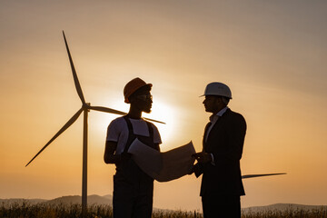 Two multiracial industrial workers examining wind turbines blueprints with amazing sunset on...