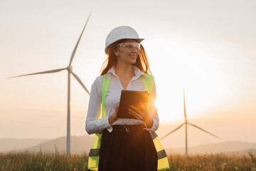 Beautiful caucasian woman in white helmet working with digital tablet at renewable energy farm. Female inspector controlling functioning of wind turbines outdoors. - obrazy, fototapety, plakaty