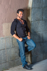 Obraz premium Dressing in a black sweater, blue jeans and leather shoes, a handsome, sexy, middle age guy with mustache and beard is leaning against the wall, smilingly looking at you..