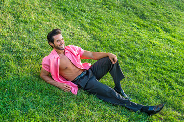 Dressing in a light red short sleeve shirt, black pants and leather shoes, a handsome, sexy, middle age guy with mustache and beard is lying on green grasses, relaxing..