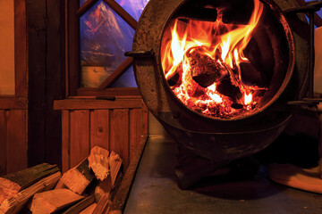 A fire is burning in the firebox of a round cast-iron stove with an open door. Firewood is lying...
