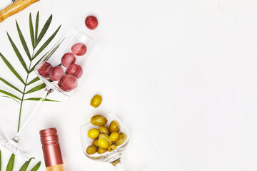 Mediterranean food composition with wine glasses with grapes and green olives, bottle of rose wine and palm leaves on white background with copy space. The concept of a summer holiday resort and relax
