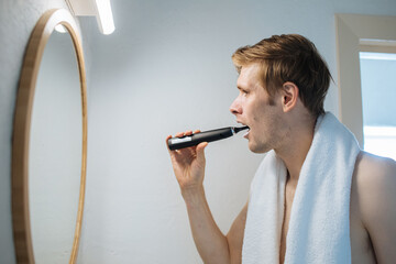 Man is using electric toothbrush. Male brushing teeth and looking in mirror. Daily dental hygiene and oral health. Rear view of young man cleaning his teeth