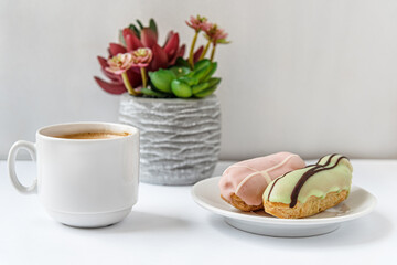 raspberry and pistachio eclairs on a porcelain plate, a cup of fresh aromatic coffee and and succulent in a pot on a white table
