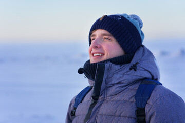 Portrait of happy cheerful positive guy, young handsome joyful man is smiling, enjoying walking outdoors in winter park at snowy frosty sunny day in warm clothes, knitted hat with backpack.