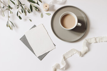 Obraz na płótnie Canvas Summer still life. Cup of coffee, blank greeting card mockup with plate, envelope. Olive tree branch and silk ribbon, white table background. Wedding stationery. Flat lay, top view. Breakfast concept