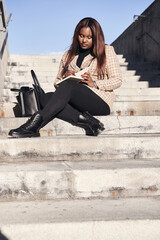 Young, African-American student sits outside on the university steps writing notes in a notebook.