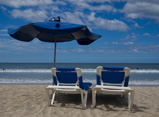 Two lounge chairs and a blue beach umbrella on a sandy beach.