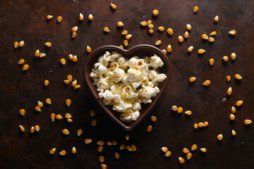 Popcorn in a brown wooden plate in the shape of a heart on a wooden table. Popcorn is laid out on the table.