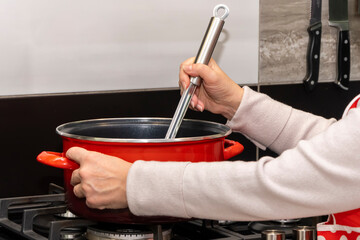 A female cook mixes food in a saucepan with a slotted spoon. The pot is on the gas stove, close-up.