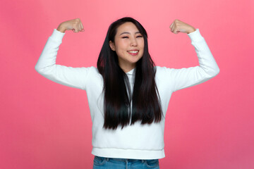 Smiling strong independent asian young girl showing muscles gesture on pink studio background. Achievements concept