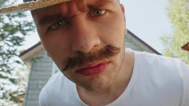 Indignant look and approaches closely into the camera of a young funny mustachioed redneck man in straw hat in front of his house. Cinematic wide angle lens closeup face portrait with distortion