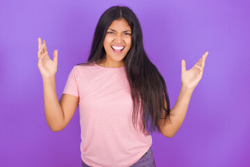 Crazy outraged Hispanic brunette girl wearing pink t-shirt over purple background screams loudly...