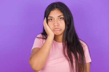 Sad lonely Hispanic brunette girl wearing pink t-shirt over purple background touches cheek with hand bites lower lip and gazes with displeasure. Bad emotions