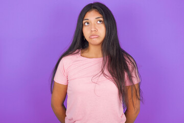 Dissatisfied Hispanic brunette girl wearing pink t-shirt over purple background purses lips and has unhappy expression looks away stands offended. Depressed frustrated model.