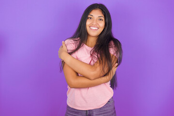 Charming pleased Hispanic brunette girl wearing pink t-shirt over purple background embraces own...