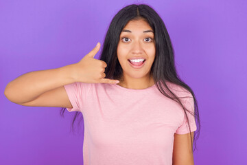 Fototapeta premium Hispanic brunette girl wearing pink t-shirt over purple background makes phone gesture, says call me back again, has glad expression.