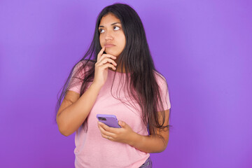 Hispanic brunette girl wearing pink t-shirt over purple background thinks deeply about something,...