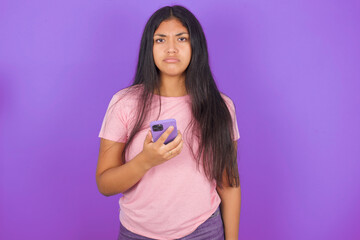 Upset dissatisfied Hispanic brunette girl wearing pink t-shirt over purple background uses mobile...
