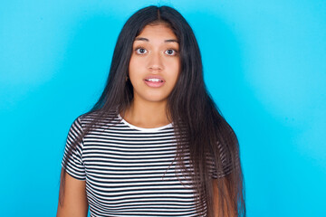 Fototapeta premium Shocked hispanic girl wearing striped t-shirt standing over blue background stares bugged eyes keeps mouth opened has surprised expression. Omg concept