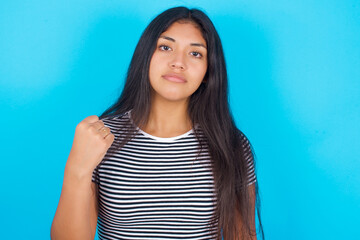 Fototapeta premium hispanic girl wearing striped t-shirt standing over blue background shows fist has annoyed face expression going to revenge or threaten someone makes serious look. I will show you who is boss