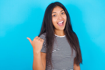 Impressed hispanic girl wearing striped t-shirt standing over blue background point back empty space