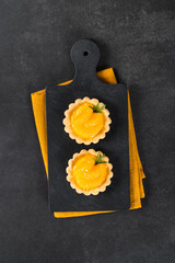 Twoo Tartlets with custard and slices of tangerines covered with syrup on a serving board on a linen napkin on a dark gray background. Top view