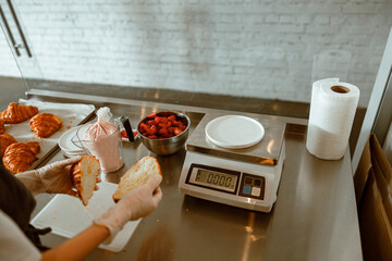 Confectioner holds cut croissant near metal table with ingredients and scales in bakery