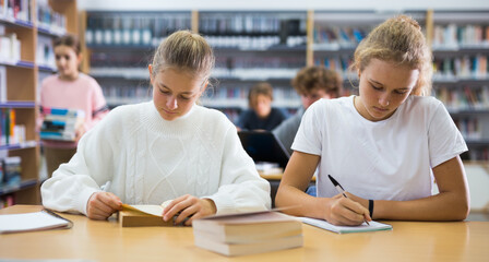 Two schoolgirls, who are engaged in lessons in the school library, write in a exercise book an...