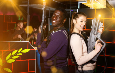 Cheerful young multiracial couple standing back to back holding laser guns during lasertag game on dark labyrinth