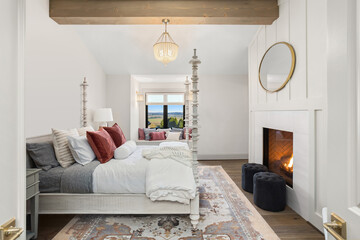 Beautiful bedroom in new traditional style luxury home with wood beam, chandelier, fireplace with roaring fire, and plush bed with elegant furnishings.