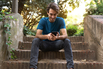 Happy young caucasian man sitting on stairs using mobile phone browsing social media in nature. Copy space.