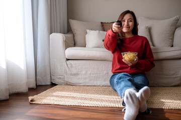 A beautiful young woman eating potato chips while searching channel with remote control to watch tv at home