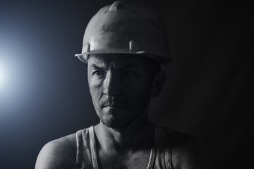 Portrait miner in helmet and undershirt with dirty face. Bright blue light on background.