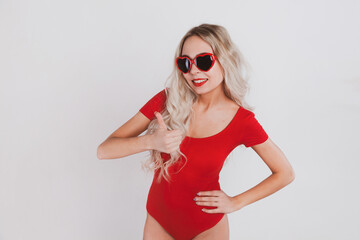 Adorable young blonde girl in red bodysuit and glasses in the shape of hearts posing on white background