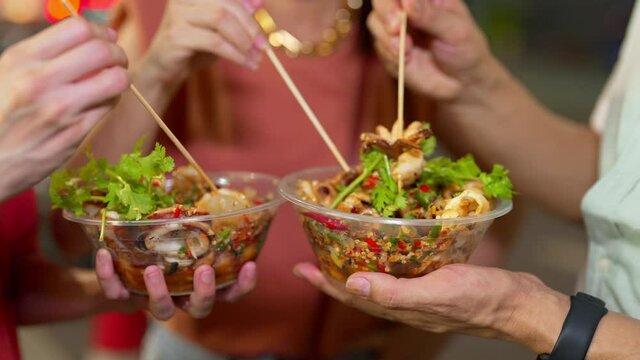 4K Group of Asian woman and LGBTQ people friends tourist enjoy eating traditional street food bbq seafood grilled squid with spicy sauce together at china town street night market in Bangkok, Thailand
