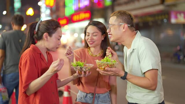 4K Group of Asian woman and LGBTQ people friends tourist enjoy eating traditional street food bbq seafood grilled squid with spicy sauce together at china town street night market in Bangkok, Thailand