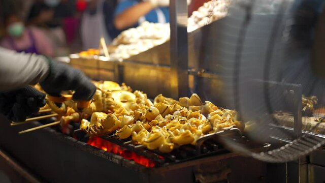 Close up of Chef hand cooking grilled squid on wooden stick with spicy sauce at street night market in Bangkok city, Thailand. Traditional delicious street food grilled seafood on charcoal grill.