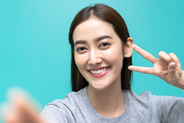Close up of beautiful young Asian woman in casual clothes taking selfie and showing gesturing v-sign, looking at camera on isolated over green pastel background. Emotions, expressions, people concept