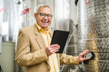 Portrait of happy senior man who owns winery. He is standing beside wine storage tanks and examining cooling process.