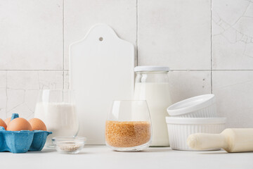 Variety of ingredients and baking utensils on white table with ceramic tile wall in the background. Kitchen background with copy space