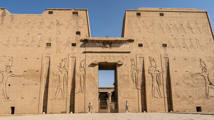 The ancient Egyptian temple of Horus in Edfu. Carved images of the gods are visible on the high wall.  There are sculptures of birds at the entrance. The columns are visible at the back of the room.
