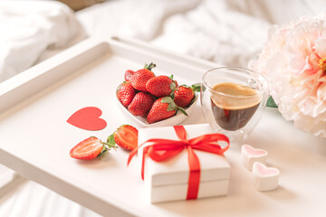 Breakfast for Valentine's Day. Heart shaped white plate with fresh strawberries, cup of coffee, gift and flowers with gift in bed. Still life composition.