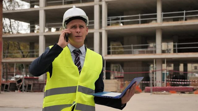 Caucasian man engineer in warnvest and hardhat standing on construction site with laptop in hands and talking on phone.