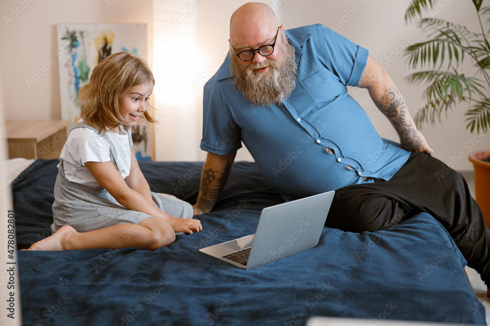 Wall mural bearded father and little girl watch moovie lesson via modern laptop sitting on bed