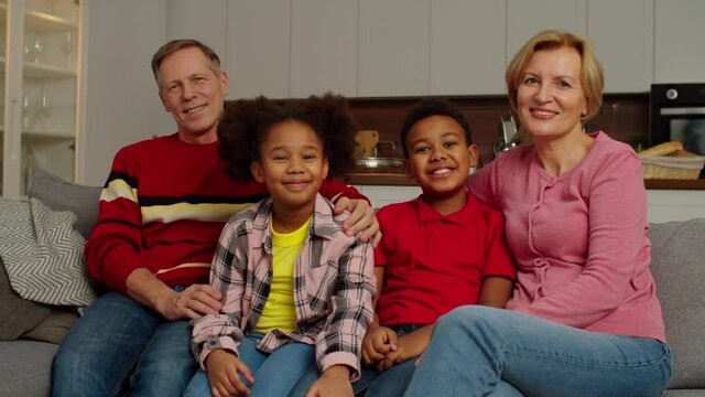 Portrait of positive loving grandparents and cute joyful elementary age multicultural children sitting on couch, looking with cheerful radiant smiles , expressing happiness and togetherness indoors.