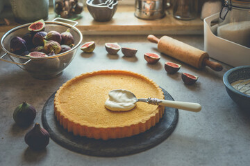 Sweet tart crust with spoon, cream and figs on kitchen table with baking tools at window