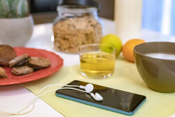 Table set for breakfast, kitchen in the background. Natural meal and technology devices. Interior view of a cozy and modern home. Millennial, youth, home working, home schooling, remote work concept.